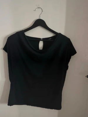 Svart topp med nyckelhålsdetalj - En svart topp med korta ärmar och rund halsringning. Toppen har en snygg nyckelhålsöppning bak som stängs med en knapp. Perfekt att styla med jeans eller kjol för en clean look. Finns bundle rabatt! Toppen har små små hål men inget som syns. 