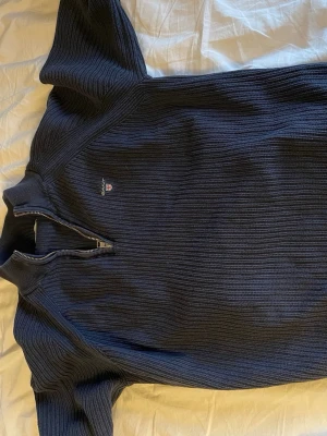 Marinblå stickad tröja från Gant - Mysig marinblå stickad tröja från Gant med ribbad struktur och half zip-dragkedja vid halsen. Klassisk logga broderad på bröstet och långärmad modell. Perfekt för lager på lager under kyliga dagar.