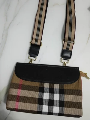 Burberry axelväska med rutigt mönster - Säljer en ny snygg axelväska från Burberry med klassiskt beige, svart, vitt och rött rutmönster. Väskan har en bred, justerbar axelrem med matchande ränder och detaljer i svart läder. Perfekt för dig som gillar ikoniska designer och vill sticka ut.