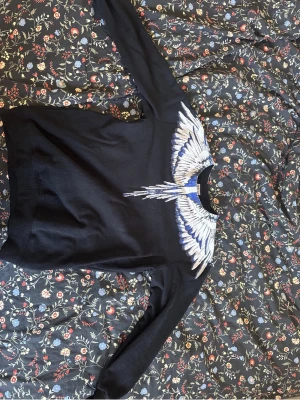 Marcelo burlon svart - Säljer en svart hoodie med ett stort, detaljerat vingtryck i vitt och blått över axlar och bröst. Hoodien har klassisk passform och ribbade muddar. Perfekt för dig som vill sticka ut med en unik design.