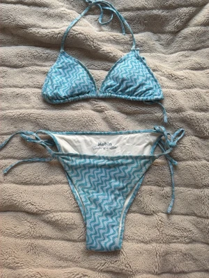 Blå glittrig bikini från Robin - Snygg blå bikini från Robin med glittrigt vågmönster. Både överdel och underdel har knytband i sidorna och klassisk triangelmodell. Perfekt för stranden eller poolen, och materialet känns mjukt och bekvämt mot huden.