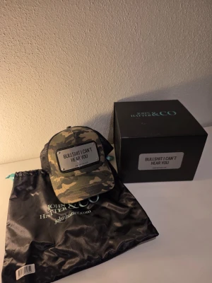 Camo keps från John Hatter & Co - Snygg camouflagemönstrad keps från John Hatter & Co med svart mesh baktill och justerbar snapback. Framtill sitter en metallplatta med texten 'BULLSHIT I CAN'T HEAR YOU' som ger kepsen en kaxig attityd. Perfekt för dig som vill sticka ut.