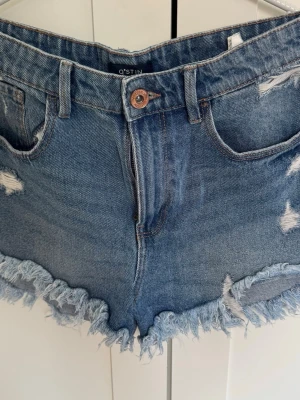 Blå fransiga jeansshorts från O'STIN - Säljer ett par blå jeansshorts från O'STIN med fransig kant och slitna detaljer. Shortsen har fem fickor, bälteshällor och en klassisk knappgylf. Perfekta för sommaren och ger en avslappnad vibe till din outfit.Skriv gärna om ni har frågor eller vill ha fler bilder!!