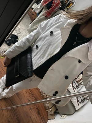 Vit trenchcoat med svarta knappar - Snygg vit trenchcoat med stora svarta knappar och klassisk krage. Jackan har en rak och kortare passform som ger en modern look. Perfekt att styla med jeans eller dressade byxor. Materialet ser ut att vara bomull eller liknande.