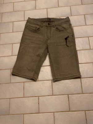 Beige denimshorts från C Denim - Säljer ett par beige denimshorts från C Denim med klassisk femficksdesign och knappstängning. Shortsens längd går till knäna och de har en diskret slitning på ena benet. Perfekta för en avslappnad och cool stil.