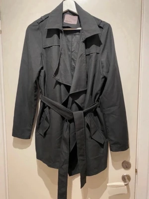 Svart trenchcoat - Svart trenchcoat med krage och knytskärp i midjan. Jackan har långa ärmar, två framfickor och ett snyggt fall framtill. 