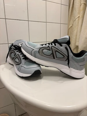 Grå och vita sneakers (pris kan diskuteras) - Snygga sneakers i grått och vitt med svarta detaljer och svarta skosnören. Skorna har en sportig design med mesh och syntetmaterial, samt en tjock vit sula som ger en modern look. Perfekta för dig som gillar stilrena och bekväma sneakers.