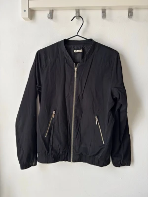 Svart bomberjacka med dragkedjor - Stilren svart bomberjacka med silverfärgade dragkedjor framtill och på fickorna. Jackan har en klassisk bomberkrage, resår vid ärmslut och nederkant samt en minimalistisk look. Perfekt för dig som gillar enkel och clean stil. Obs: Det finns hål på insidan av fickorna, men det är inget som syns utåt och går enkelt att sy.
