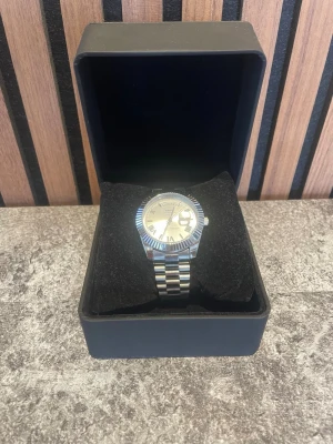 Silverfärgad klocka från Nibosi - Stilren klocka från Nibosi i silverfärg med klassisk länkarmband och räfflad bezel. Urtavlan är ljus med romerska siffror, datum- och veckodagsvisning. Klockan har en elegant och tidlös design som passar till många olika stilar.