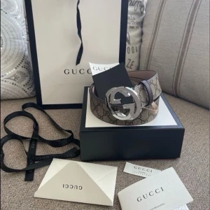 Gucci beige GG-bälte med silverdetalj - Säljer ett ikoniskt Gucci-bälte i beige med det klassiska GG-mönstret och en stor, rund silverfärgad logga som spänne. Bältet är gjort i skinn och har en lyxig känsla som lyfter vilken outfit som helst.