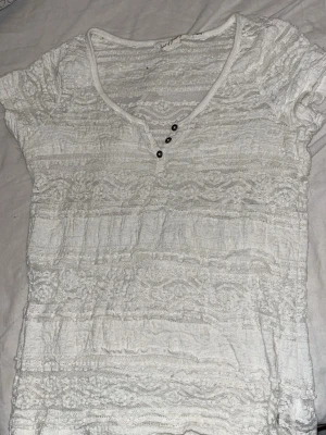 Vit spetsblus med knappar - Säljer en vit blus i spets med v-ringning och tre dekorativa knappar framtill. Toppen har korta ärmar och är figurnära med fina spetsdetaljer i hela tyget. Perfekt för dig som gillar romantisk och feminin stil.