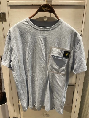 Ljusblå t-shirt med ficka från Lyle & Scott - Snygg ljusblå t-shirt från Lyle & Scott med klassisk passform och en stor bröstficka med logga. T-shirten har rund halsringning och är tillverkad i mjuk bomull, perfekt för en avslappnad stil.