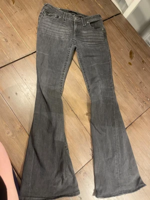 Grå bootcut jeans från True Religion - trasiga  - Trasiga. De har ett hål i skrevet som behöver lagas. Se bild. Säljer ett par grå bootcut jeans från True Religion med klassiska fickor bak och snygga detaljer vid ficklocken. Jeansen har låg midja, bälteshällor och är tillverkade i denim med lätt stretch. Perfekta för dig som gillar en avslappnad men trendig look.