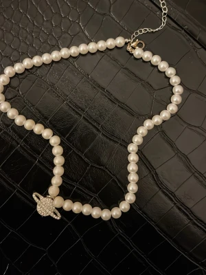 Pärlhalsband med planet-hänge - Snyggt halsband med vita pärlor och ett glittrande planetformat hänge framtill. Halsbandet har en justerbar kedja i silverfärg och pärlorna är jämnt runda. Perfekt för dig som gillar unika detaljer och vill sticka ut.