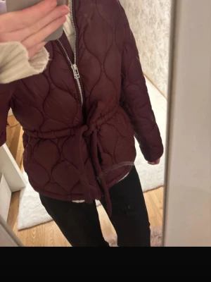 Vinröd quiltad pufferjacka med knytband - Snygg vinröd pufferjacka med quiltat mönster och dragkedja framtill. Jackan har ett knytband i midjan för en mer markerad siluett och är perfekt för kalla vinterdagar. Materialet är vadderat och ger en mysig känsla. Inga synliga loggor eller märken.