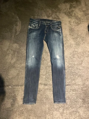 Blå slim fit jeans från Replay - Snygga blå slim fit jeans från Replay med slitningar på knäna och ljusa tvättade partier. Klassisk femficksmodell med detaljerade sömmar och logotyp på myntfickan. Perfekta för en trendig och avslappnad stil.