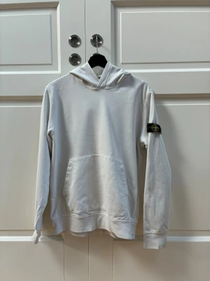 Vit hoodie från Stone Island - Säljer en clean vit hoodie från Stone Island med klassisk patch på vänster ärm. Hoodien har känguruficka framtill, ribbade muddar och en skön huva. Perfekt för dig som gillar streetwear och vill ha en enkel men snygg look. Materialet känns mjukt och bekvämt. Tröjan har tyvärr några fläckar men många av dem syns inte på håll.