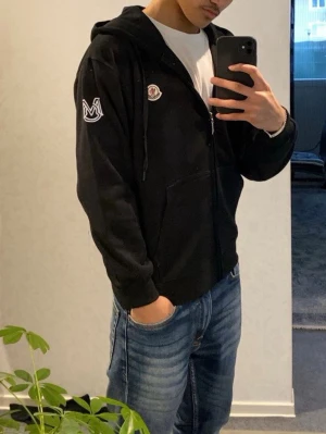 Svart Moncler hoodie med dragkedja - Svart hoodie från Moncler med dragkedja framtill, huva med snören och två fickor. Ikonisk Moncler-logga på bröstet och ett vitt broderat M-märke på ärmen. Tillverkad i mjukt bomullsmaterial som känns skönt mot huden. Perfekt för en chill och stilren look. INTE ÄKTA!