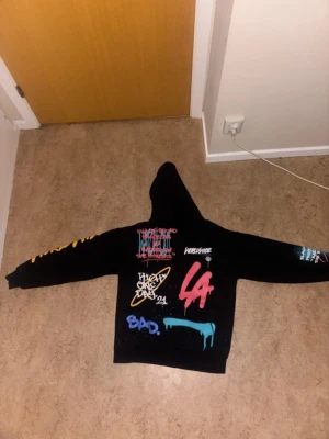 Svart hoodie med färgglad graffiti - Säljer en svart hoodie med huva och färgglada graffiti-tryck över hela plagget. Trycken inkluderar ord som 'BAD', 'DRIP2HARD', 'LA' och olika motiv i blått, rött, gult och vitt. Hoodien har en avslappnad passform och streetwear-vibe.