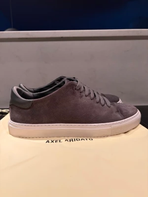 Gråa Axel Arigato sneakers i mocka - Stilrena grå sneakers från Axel Arigato som är använd 2-3 gånger!i mjuk mocka med matchande grå skosnören och vit sula. Klassisk låg modell med minimalistisk design och diskreta detaljer. Perfekta för dig som gillar en clean och modern look.
