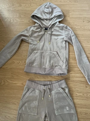 Beige velourset från Juicy Couture - Säljer ett beige velourset från Juicy Couture i storlek XS. Setet består av en ziphoodie med fickor och guldfärgade detaljer samt matchande byxor med dragsko och fickor. Perfekt för chill dagar hemma eller när du vill vara extra comfy.
