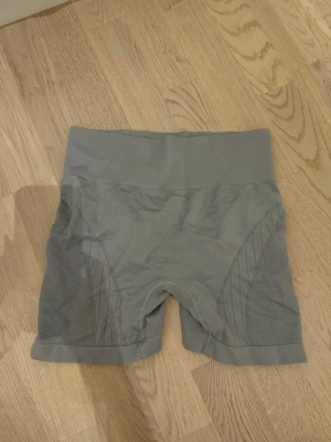 Grå  träningsshorts - Gråa  shorts med hög midja och ribbad linning. Tillverkade i stretchigt syntetmaterial som andas, perfekta för träning eller gym. Snygga meshdetaljer längs sidorna för extra ventilation och stil.