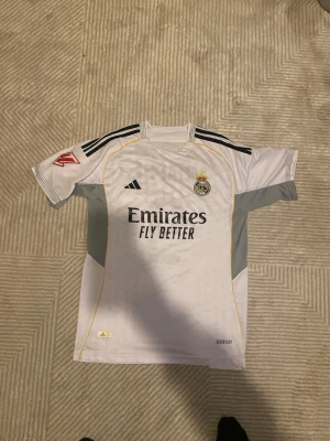 Real Madrid fotbollströja Mbappé - Säljer en vit Real Madrid fotbollströja från Adidas med Mbappé och nummer 9 på ryggen. Tröjan har svarta detaljer, klubbmärke på bröstet och sponsortext framtill. Perfekt för fotbollsfans som vill sticka ut på planen eller läktaren.