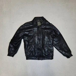 Vintage Black Leather Jacket – 100% Real Leather  - Klassisk svart skinnjacka i 100% äkta läder. Kort modell med dragkedja, två fickor fram och elastisk nederkant/ärmslut. Insidan har ett coolt mönstrat foder med guldtryck (typ vintage-karta/stadsmotiv). Storlek: S – sitter som en liten M (bra för någon runt 170–175 cm). Skick: Mycket bra skick – knappt använd (köpt från vintagebutik för 4000 kr). Inga defekter, inga stora slitage eller repor. Läderet är mjukt och fint. Säljer för att den är lite för stor på mig (jag är 172 cm). Packas säkert 