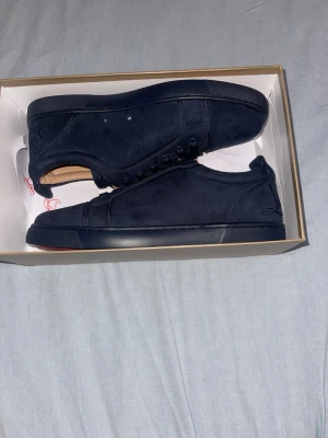 Mörkblåa sneakers från Christian Louboutin - Snygga mörkblå sneakers från Christian Louboutin i mocka med klassisk röd sula. Skorna har en stilren design med ton-i-ton snörning och diskret logga på hälen. Perfekta för dig som vill ha en clean look med exklusiv känsla.