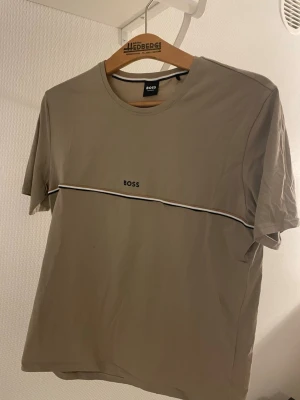 Beige t-shirt från BOSS med rand - Snygg beige t-shirt från BOSS med svart och vit rand tvärs över bröstet och diskret BOSS-logga. Klassisk rund halsringning och avslappnad passform. Perfekt för en stilren och enkel look.