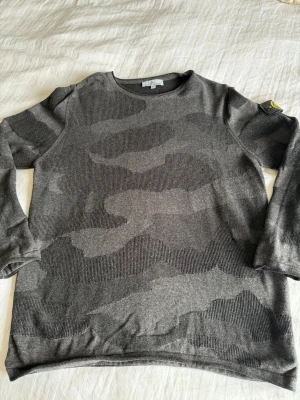 Grå camo-mönstrad tröja Stone Island - Snygg grå långärmad tröja från Stone Island med diskret camo-mönster och klassisk patch på ärmen. Tröjan har rund halsringning och är tillverkad i mjukt material som känns skönt mot huden. Perfekt för dig som gillar streetwear och stilrena detaljer. Passar mer en xl 