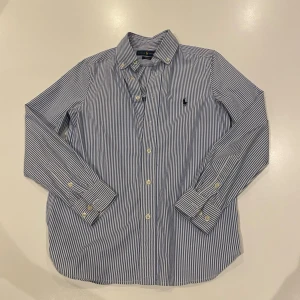 Randig vit blå skjorta från Ralph Lauren - Klassisk blåvit randig skjorta från Ralph Lauren med knappar framtill och lång ärm. Skjortan har en broderad logga på bröstet och är tillverkad i bomull. Perfekt för en stilren look.