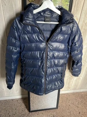 Mörkblå dunjacka från Sail Racing - Snygg mörkblå dunjacka från peak performance med glansig finish, huva och dragkedja framtill. Jackan har två sidofickor med dragkedja och logga på ärmen. Perfekt för kalla vinterdagar och riktigt skön att bära.