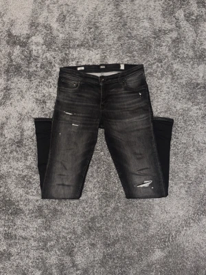 Svarta slitna jeans från Jack & Jones - Snygga svarta jeans från Jack & Jones med slim-fit passform och slitningar på benen. Jeansen har fem fickor, dragkedjegylf och en tvättad look med gråa nyanser. Perfekta för en trendig streetstil.