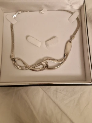 Silverfärgat halsband Fifth Avenue - Snyggt silverfärgat halsband från Fifth Avenue Collection med en elegant infinity-formad detalj framtill. Ena sidan av hänget är dekorerad med små gnistrande pärlor som ger extra bling. Perfekt accessoar för att lyfta din outfit. Helt ny och kommer med låda och allt. Kostar ca 60 dollar ny, men går inte att köpa på deras websida!