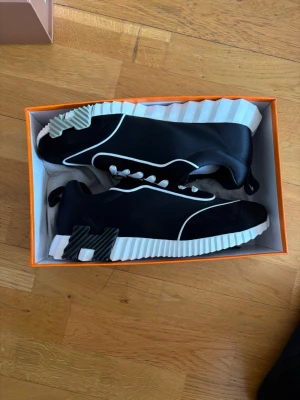 Svarta sneakers med vit sula - Snygga svarta sneakers med vit räfflad sula och vita detaljer längs kanterna. Skorna har en unik design med kontrasterande material och en cool H-detalj på hälen. Perfekta för dig som vill sticka ut med exklusiva sneakers.