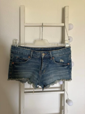 Blå denimshorts med låg midja - Säljer ett par klassiska blå denimshorts med fransig kant och slitningar. Shortsen har fem fickor, knapp och dragkedja framtill samt låg midja. Perfekta för varma dagar och en avslappnad look. Är lågmidjade!