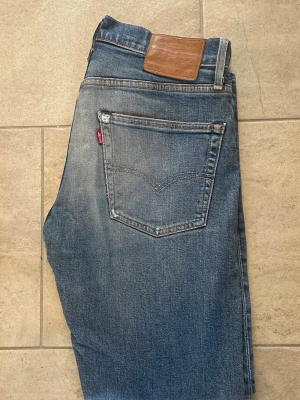 Levi's blå jeans med raka ben - Säljer ett par klassiska blå Levi's jeans med raka ben och femficksdesign. Jeansen har en snygg slitning på framsidan och en brun läderpatch bak i midjan. Materialet är robust denim och passformen är tidlös. Perfekt för dig som gillar ikoniska jeans.