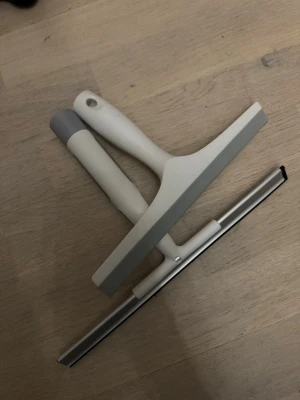 Två fönsterskrapor i plast och metall - Säljer två praktiska fönsterskrapor / duschskrapor, en i vit plast med grått handtag och en i metall med svart gummilist. Perfekta för att hålla speglar och fönster rena. Ergonomiska handtag för bra grepp och enkel användning.