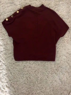 Vinröd topp med guld knappar på axeln - Säljer en vinröd topp med korta ärmar och dekorativa guldfärgade knappar längs ena axeln. Toppen har en högre halsringning och är i ett mjukt, stretchigt material. Perfekt för dig som vill ha en enkel men ändå unik look. Tröjan är köpt på Zara 2024 och är i bra skick!