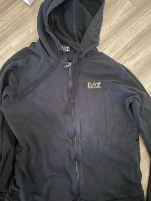 Svart EA7 hoodie med dragkedja - Säljer en svart hoodie från EA7 Emporio Armani med dragkedja framtill och justerbar huva. Hoodien har guldfärgad logga på bröstet och dekorativa guldstreck på ena ärmen. Perfekt för en chill och stilren look.