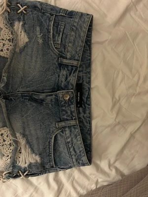 Blå jeansshorts med spets och snörning - Säljer ett par blå jeansshorts från tally wejlimed slitna detaljer, spetsinslag och snörning i sidorna. Shortsen har fem fickor, bälteshällor och knappgylf. Perfekta för sommaren och ger en cool, avslappnad vibe.