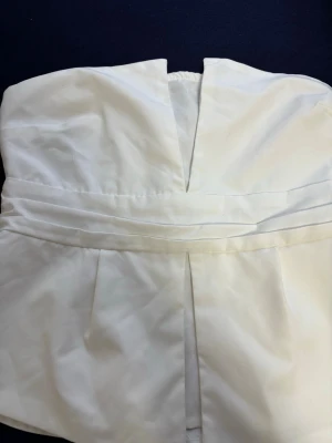 Vit axelbandslös tubtopp med v-ringning - Snygg vit axelbandslös tubtopp med markerad midja och djup v-ringning framtill. Toppen har veckade detaljer och dragkedja i ryggen för enkel påklädning. Perfekt för att skapa en clean och stilren look.