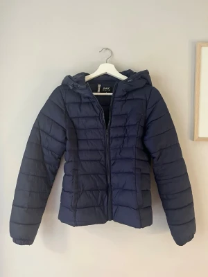 Mörkblå pufferjacka från ONLY - Snygg mörkblå pufferjacka från ONLY med huva och dragkedja framtill. Jackan har quiltad design, två sidofickor och elastiska muddar vid ärmsluten. Perfekt för kalla dagar och enkel att matcha med olika outfits.
