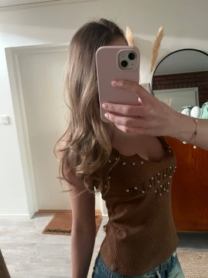 Brun ribbad linnetopp med pärlor - Säljer en brun ribbad linnetopp med pärlor på bröstet. Toppen har en v-ringad form och är ärmlös, perfekt för att skapa en snygg och avslappnad look. Materialet känns mjukt och stretchigt, och toppen har en croppad passform.