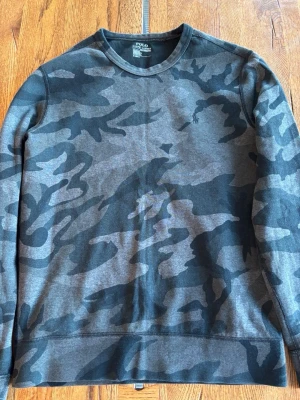 Camo crewneck från Polo Ralph Lauren - Snygg grå och svart camo-mönstrad crewneck från Polo Ralph Lauren. Tröjan har rund halsringning och diskret broderad logga på bröstet. Perfekt för dig som gillar streetstyle och vill ha något unikt i garderoben.