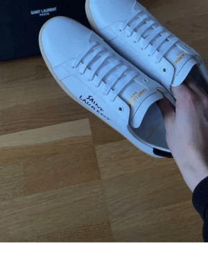 Vita Saint Laurent sneakers - Snygga vita sneakers från Saint Lauren med handskriven Saint Laurent-text på sidan och är tillverkade i slätt läder med klassisk låg profil och vita snören. Storlek 41/42/43 finns