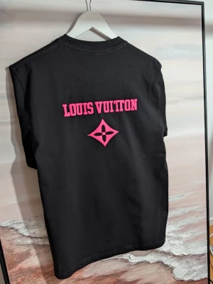 Iouis Vuitton T-shirt  - Exklusiv svart t-shirt med neonrosa 3D-logotyp Stilren t-shirt i högkvalitativ svart bomull med iögonfallande detaljer. Plagget har en centrerad logotyp på bröstet och ett större texttryck på ryggen, båda utförda i ett upphöjt, gummerat 3D-tryck i klarrosa neon. Den taktila effekten ger tröjan en modern och lyxig känsla. Färg: Svart med neonrosa detaljer. Design: Rundhalsad med korta ärmar och klassisk passform. Detalj: Markant 3D-textur på alla tryck.( Fast pris)