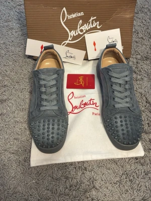 Gråa Christian Louboutin sneakers - Snygga gråa sneakers från Christian Louboutin med klassiska röda sulor och nitar på tån. Skorna är låga och har en ovandel i mocka med matchande grå skosnören. Perfekta för dig som vill sticka ut med exklusiv design och coola detaljer.