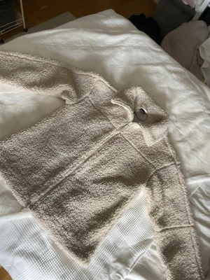 Beige teddyjacka med dragkedja - Mysig beige teddyjacka med hög krage och hel dragkedja framtill. Jackan har en fluffig och mjuk struktur som ger en avslappnad look. Perfekt för kyliga dagar och enkel att matcha med olika outfits.